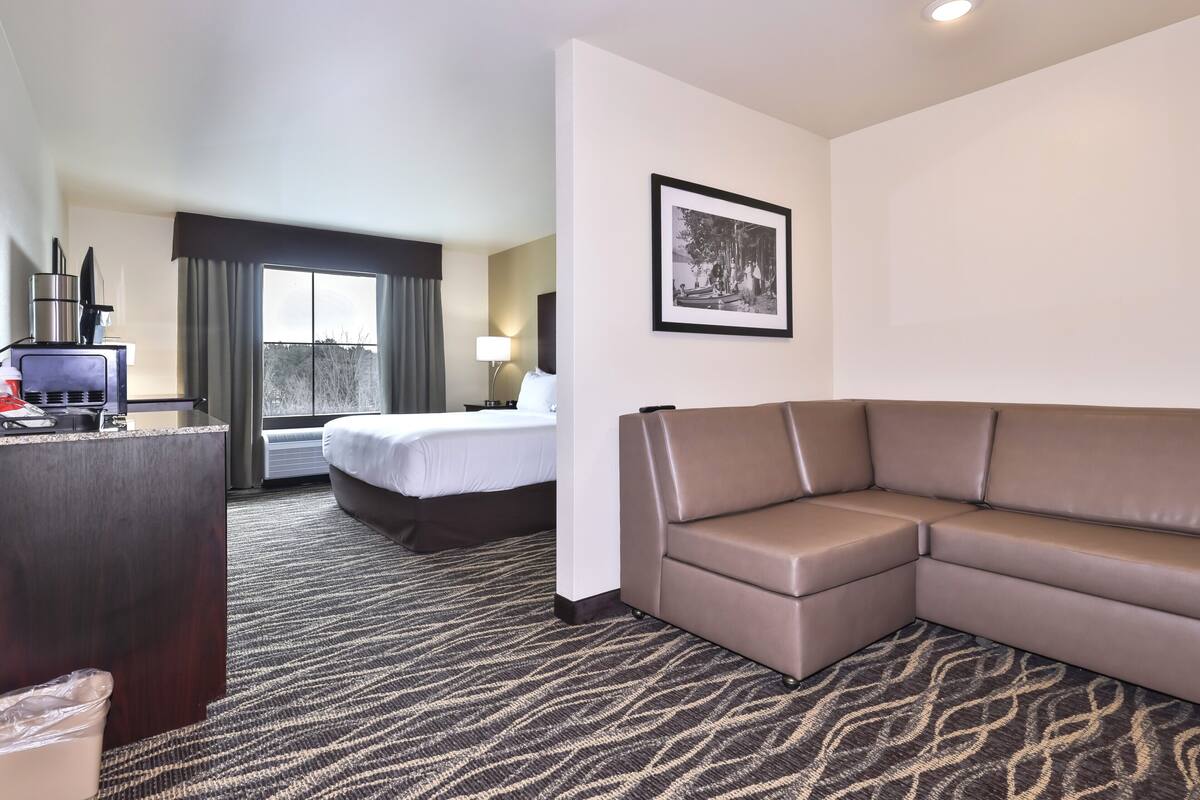 Habitación de hotel con cama, sofá, escritorio con computadora, ventana con cortinas y un cuadro en la pared.