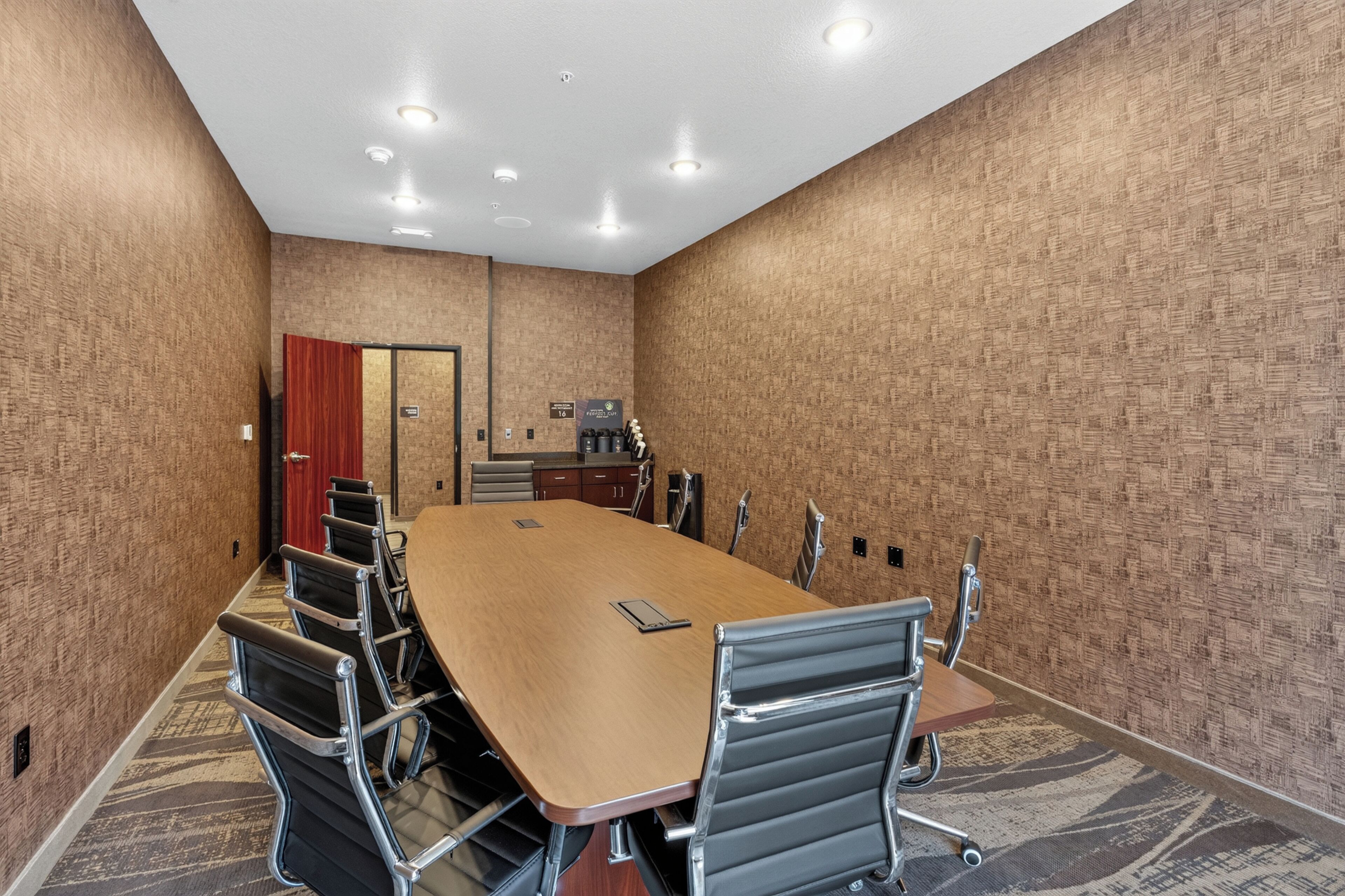 A conference room with a long table, chairs, a red door, a kitchenette, and a patterned carpet.