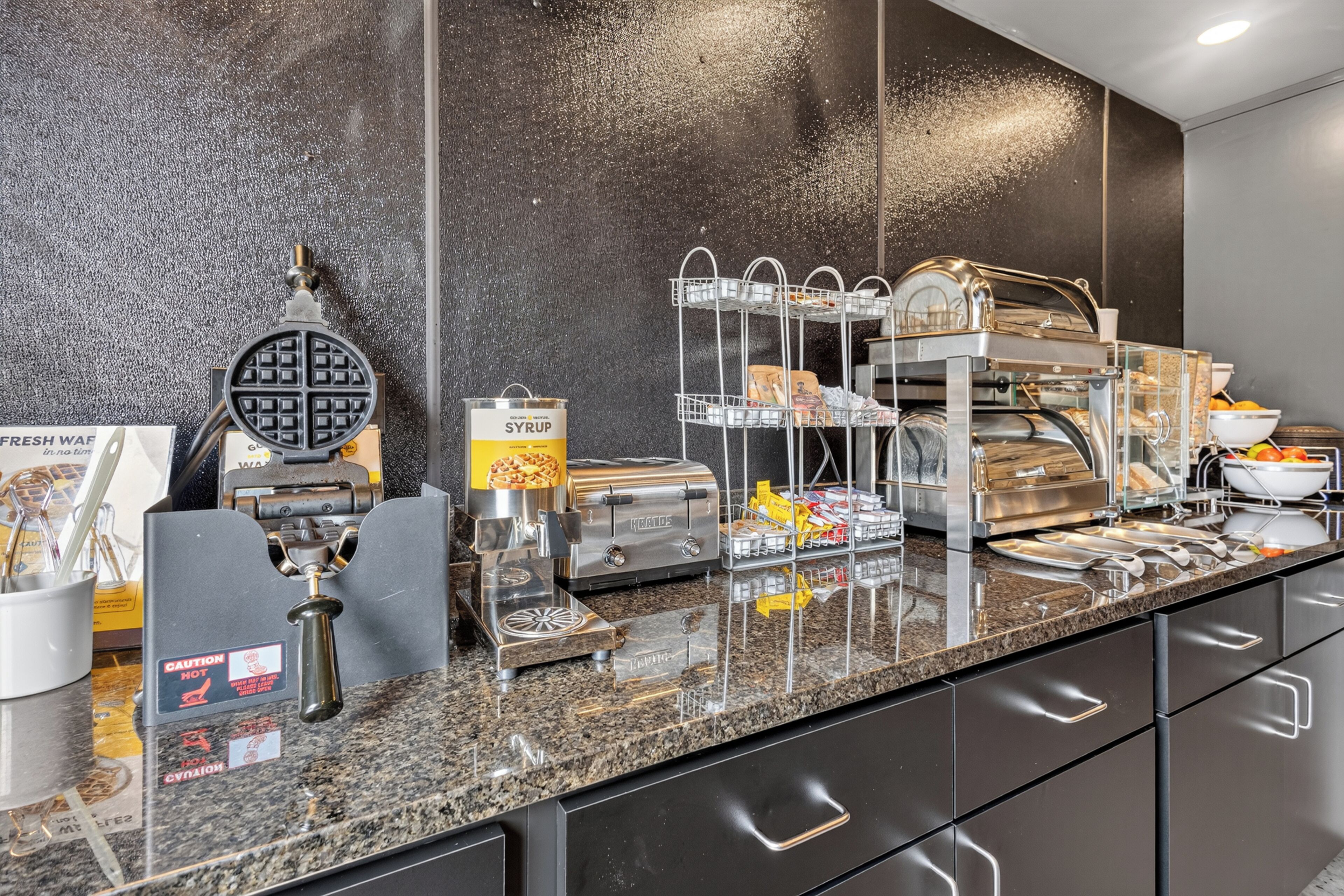 A breakfast buffet area with a waffle maker, syrup dispenser, and various food items.