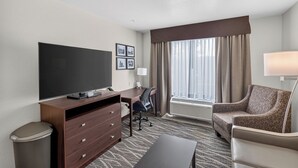 A hotel room with a flat-screen TV, a desk with a chair, a sofa, a waste bin, and a large window with curtains. - Cobblestone Hotel & Suites - Waukesha/West Milwaukee (Waukesha)