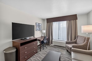 A hotel room with a flat-screen TV, a desk with a chair, a sofa, a waste bin, and a large window with curtains. - Cobblestone Hotel & Suites - Waukesha/West Milwaukee (Waukesha)
