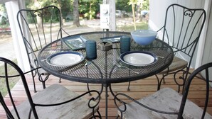 A round metal table with two plates and a blue bowl, surrounded by metal chairs on a wooden deck.
