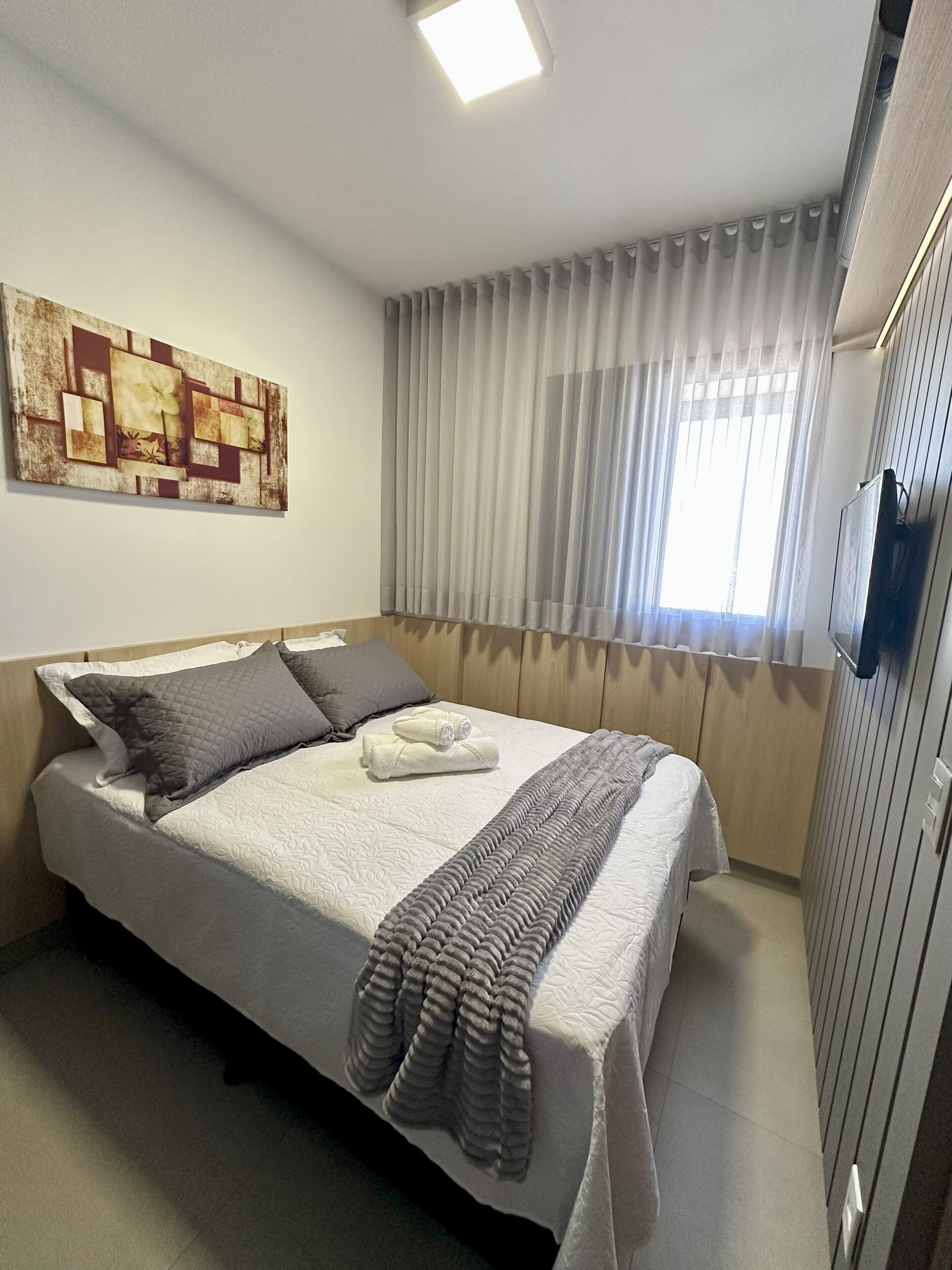 Una cama bien hecha con almohadas, una manta y una toalla doblada. Hay un cuadro en la pared y una ventana con cortinas.