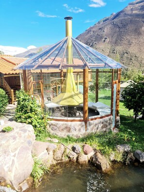 A rustic wooden gazebo with a glass roof, a stone fire pit, and a small stream in the foreground.
