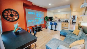 A modern living room with a large screen displaying a video game, a dining area with a black table, and a kitchen in the background.