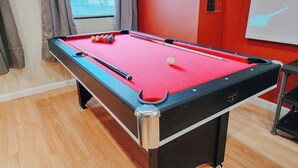 A pool table with balls and cues, a window with curtains, and a projector screen on the wall.