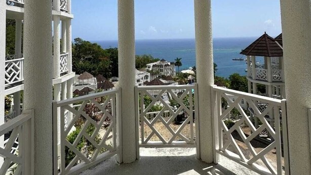 A covered outdoor area with white columns and railings, overlooking a coastal view with buildings and the sea. - Charming 1-bedroom apartment with Breathtaking views in Ocho Rios, Jamaica (Ocho Rios)