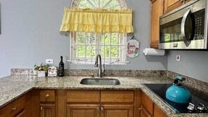 A kitchen with wooden cabinets, a granite countertop, a stainless steel microwave, a blue kettle, and a window with a yellow valance. - Charming 1-bedroom apartment with Breathtaking views in Ocho Rios, Jamaica (Ocho Rios)