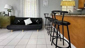 A living room with a black sofa, a coffee table, a lamp, and a bar area with stools. There is a window with a yellow curtain and a sign on the wall. - Charming 1-bedroom apartment with Breathtaking views in Ocho Rios, Jamaica (Ocho Rios)