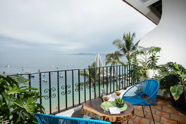 A balcony with a view of the sea, a table with a drink, and blue chairs.