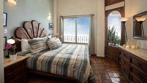 A bedroom with a large bed, a dresser, a nightstand, a potted plant, and a view of the sky through a window.
