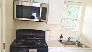A compact kitchen with a black stove, white cabinets, a stainless steel microwave, and a window with blinds. - Inclinescape#4 (Colorado Springs)