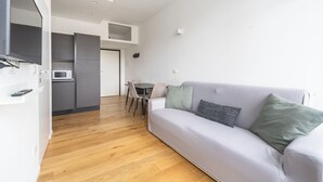 Ett modernt vardagsrum med en grå soffa, ett matbord med stolar, en köksdel med mikrovågsugn och ett trägolv.