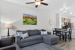 A modern living room with a gray sectional sofa, a dining table with chairs, a ceiling fan, a kitchen area, and a landscape painting on the wall. - Brownstone Beauty - Boise, ID (Southwest Ada County Alliance)