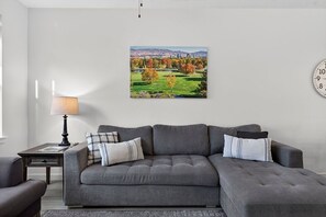 A living room with a grey sofa, a lamp, a clock, and a cityscape painting on the wall. - Brownstone Beauty - Boise, ID (Southwest Ada County Alliance)