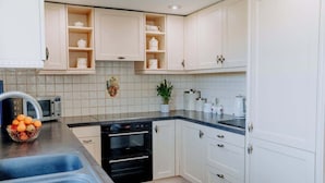 A modern kitchen with white cabinets, a black oven, and a countertop with a bowl of fruit. - Charming Rural Escape with Play Room BBQ (Llawhaden)