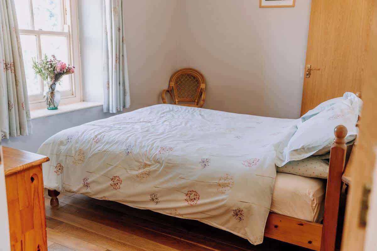 Une chambre à coucher avec un lit en bois, une chaise en osier, une commode en bois et une fenêtre avec des rideaux.