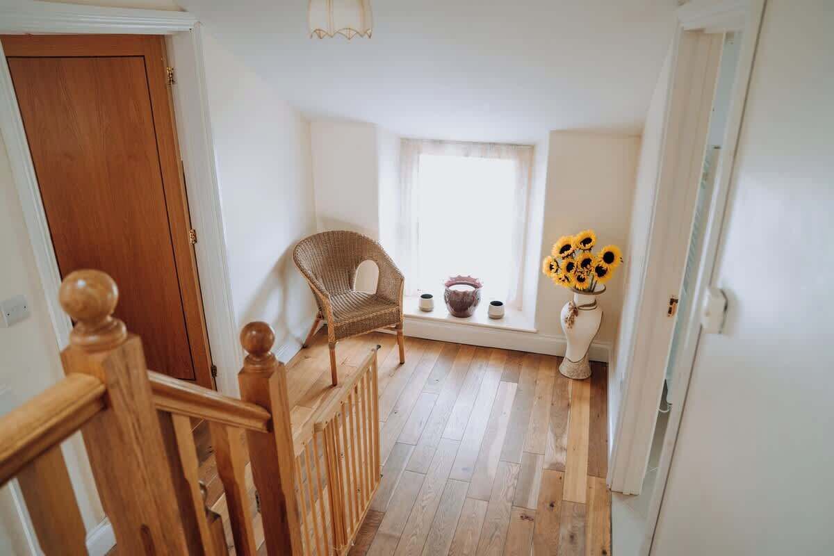 Una escala condueix a una habitació amb una cadira de vimbles, una taula petita amb un ram de girassols i una finestra amb cortines translúcides.
