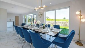 Un coin repas avec une longue table dressée pour six personnes, des chaises bleues, un lustre et une vue sur un mur de pierre et la verdure à l’extérieur.