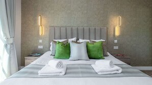 Un lit bien fait, avec du linge de lit blanc et des coussins verts, une tête de lit grise et deux tables de chevet avec des lampes.