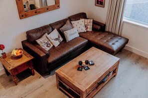 A living room with a brown leather sofa, wooden coffee table, and a small wooden side table with a bowl of fruit. - Stylish Country Escape | Scenic Views & EV Charger (Llanarthney)