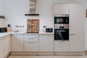A modern kitchen with white cabinetry, a stainless steel range hood, a microwave, and an oven. - Stylish Country Escape | Scenic Views & EV Charger (Llanarthney)