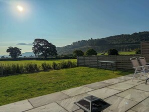 A patio area with a table and chairs, a grassy lawn, and a view of a hilly landscape. - Stylish Country Escape | Scenic Views & EV Charger (Llanarthney)