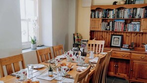 Un comedor con una mesa de madera lista para el desayuno, rodeada de sillas. A la derecha, una estantería llena de libros.