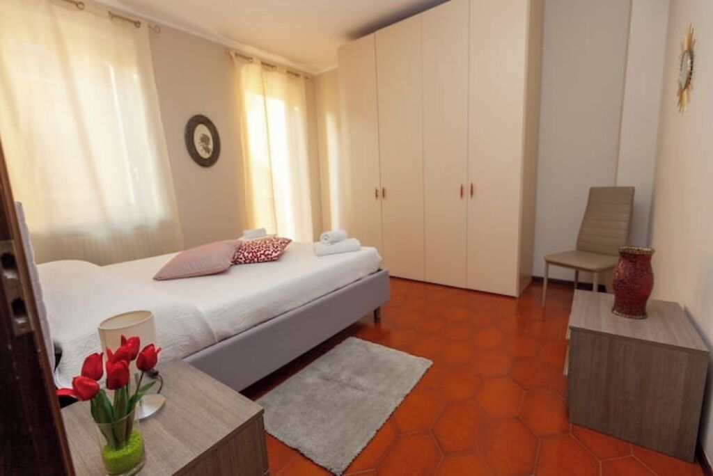 Una camera da letto con un letto, un comodino con un vaso di fiori rossi, una sedia e un'ampia armadio.