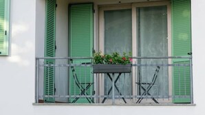Un balcón con contraventanas verdes, barandilla de metal y una maceta con flores.