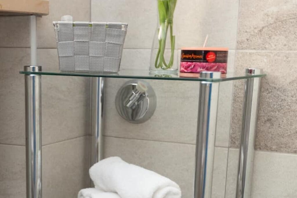 Une étagère en verre avec un panier en métal, un vase avec des fleurs vertes et une boîte de produits de toilette.