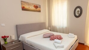Una cama bien hecha con almohadas y toallas, una mesita de noche con lámpara y un florero, y un cuadro en la pared.