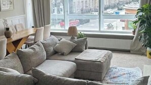 Un salon moderne avec un canapé d’angle gris, une table à manger avec des chaises et de grandes fenêtres donnant sur la ville.