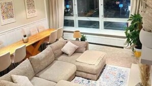 Un salon comprenant un grand canapé d’angle beige, une table à manger en bois avec des chaises, et une grande fenêtre avec des rideaux.