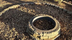 Un foyer circulaire en pierre, entouré d’une zone recouverte de gravier, avec des arbres et une étendue d’eau en arrière-plan.
