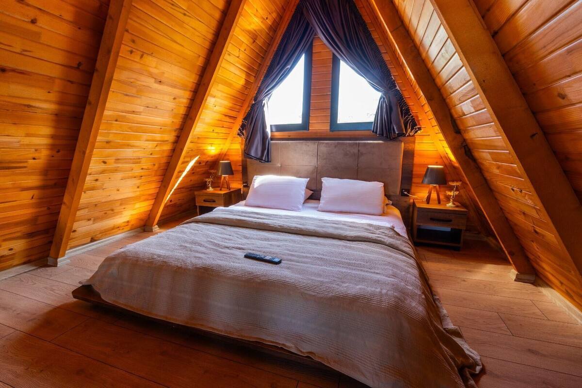 A cozy attic bedroom with a wooden sloped ceiling, a large bed with white linens, two bedside tables with lamps, and a window with curtains.