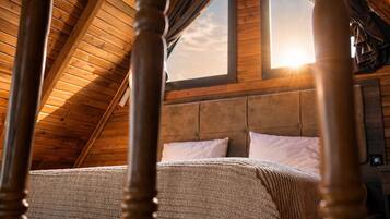A bedroom with a sloped ceiling, wooden beams, a bed with a patterned bedspread, and a window with sunlight streaming in.