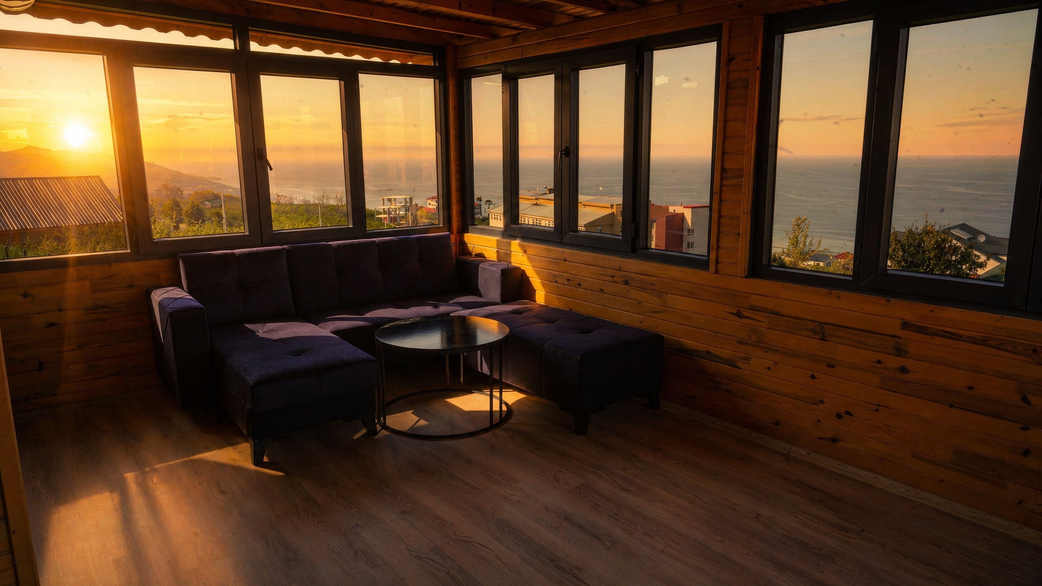 Una habitación con interior de madera, un sofá, una mesita y ventanas amplias con vistas al mar y edificios.