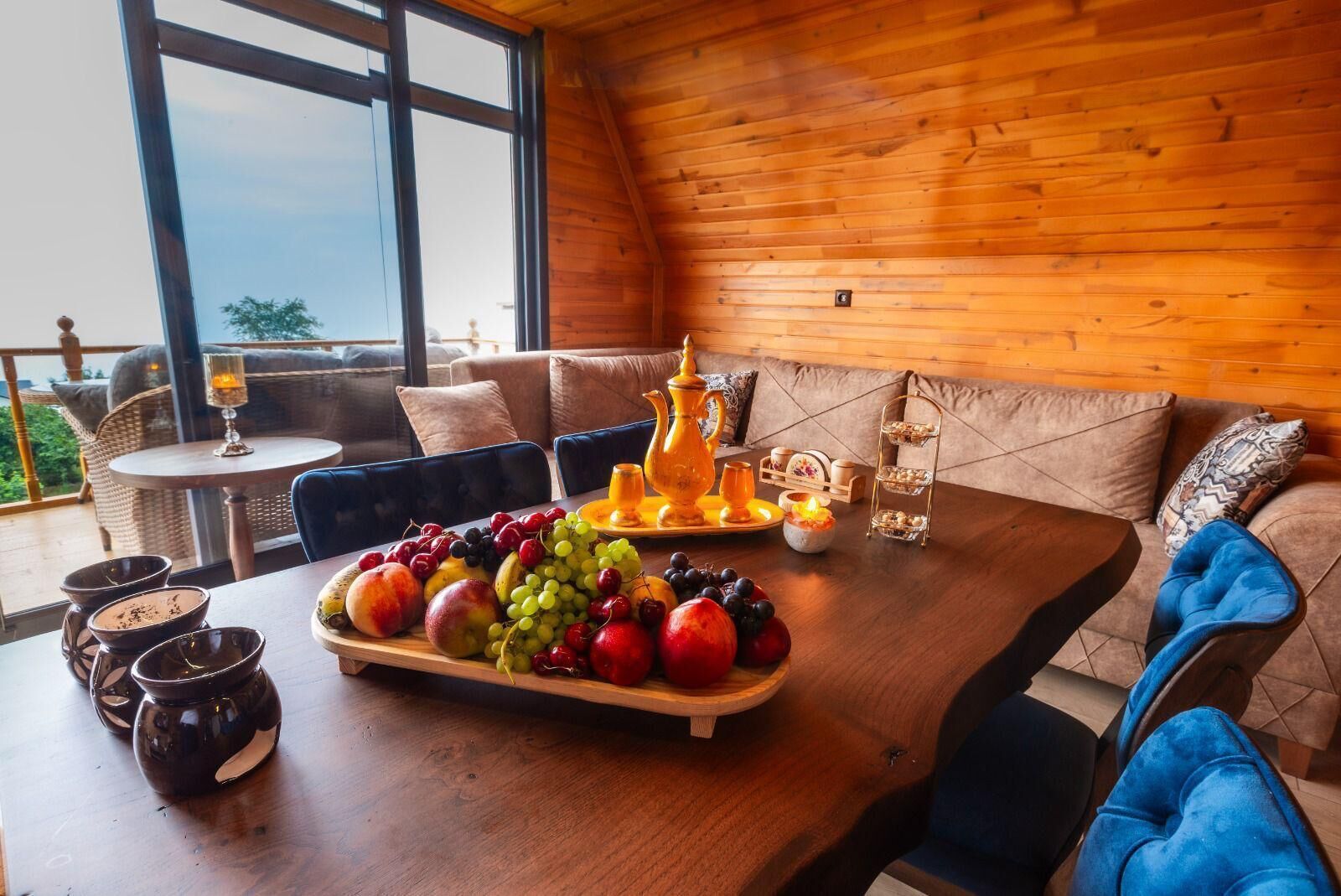 A wooden interior with a dining table set with fruits, a pitcher, and a tray. There is a sofa with cushions and a small table with a candle and a glass.