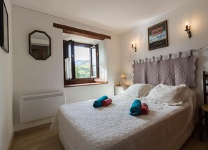 A bedroom with a large bed, a window with curtains, a wall-mounted mirror, a bedside table with a lamp, and a poster on the wall.