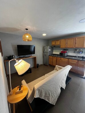 A compact room with a bed, a desk with a lamp, a television, a kitchenette with wooden cabinets, and a ceiling light.