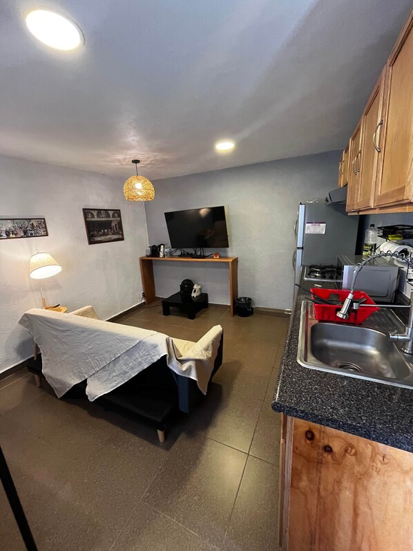 A compact living space with a bed, a TV, a small table, a kitchen area with a sink, and a dining table.