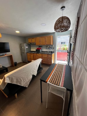 A compact room with a bed, a small table, a TV, a kitchenette, and a view of the outside.