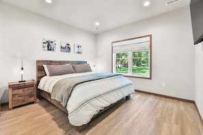 A bedroom with a wooden bed, a nightstand, and a window with a view of greenery.