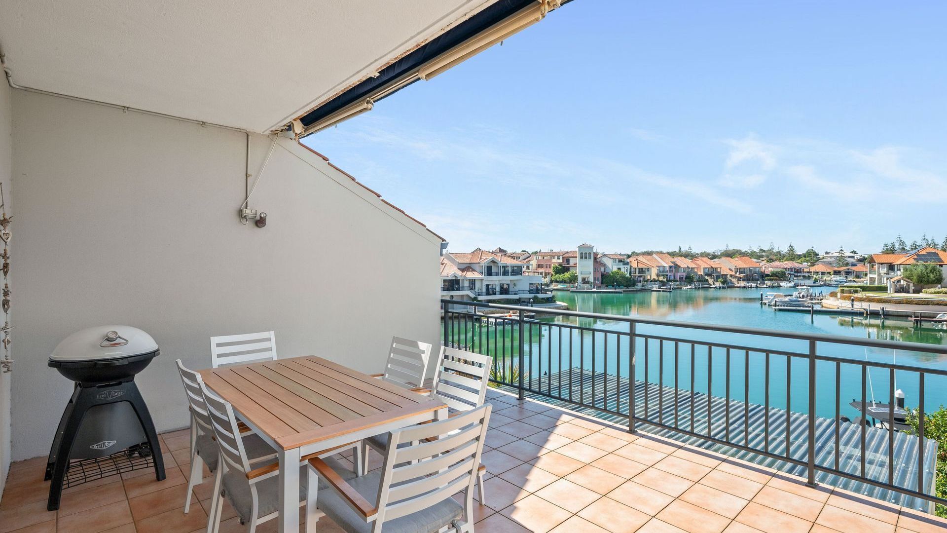 Un balcon avec une table et des chaises en bois, donnant sur une marina où l’on peut voir des bateaux et des maisons.