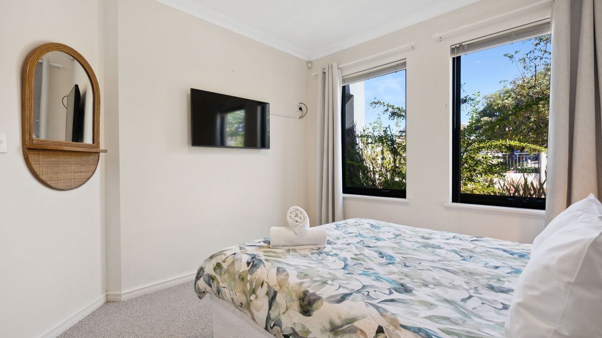 Un dormitorio con cama, televisor de pantalla plana, espejo redondo y ventanas amplias que dan a vistas de vegetación.