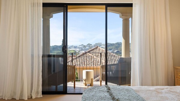 A bedroom with a large sliding glass door leading to a balcony with a view of a tiled roof and distant buildings.