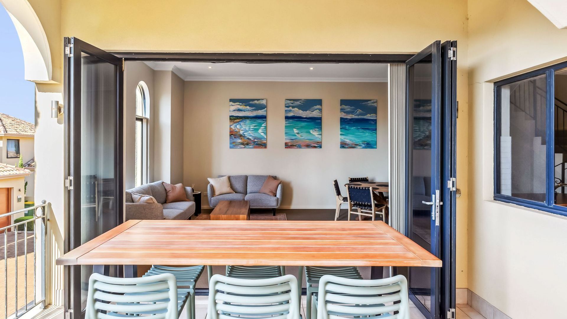 An open patio with a wooden table and chairs, a sofa, and a dining table with chairs. There are three paintings on the wall depicting ocean scenes.