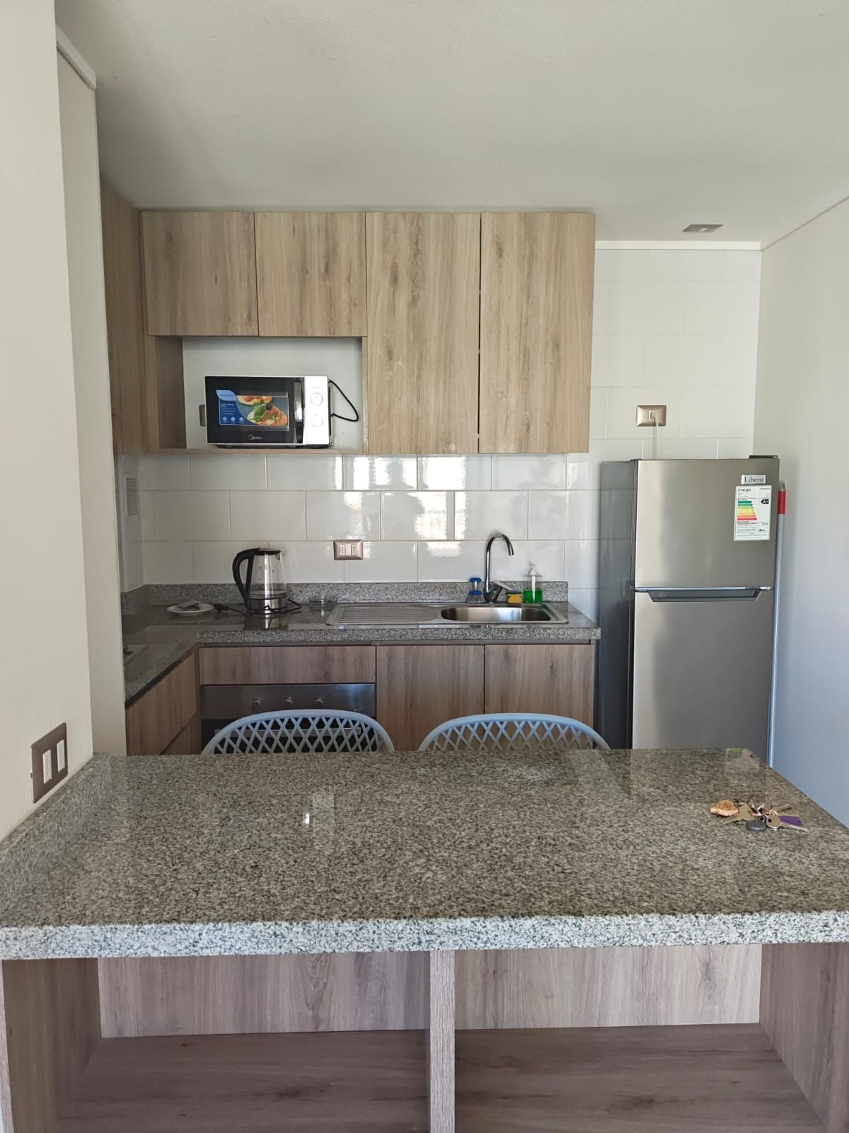Una cocina compacta con armarios de madera, un refrigerador de acero inoxidable, un microondas y una encimera de granito.