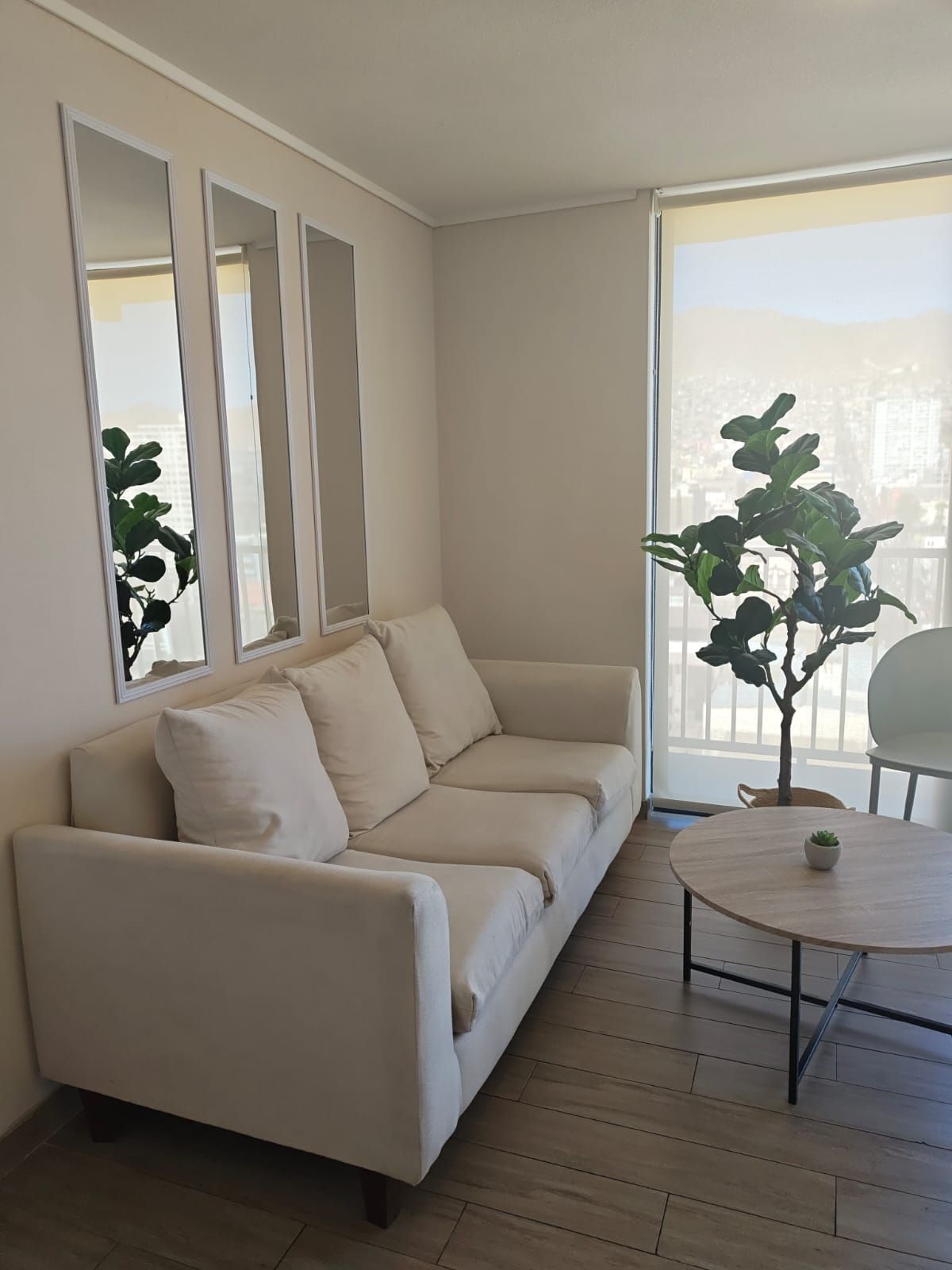 Uma sala de estar moderna com um sofá bege, uma mesa redonda de madeira e uma janela ampla com vista para edifícios.